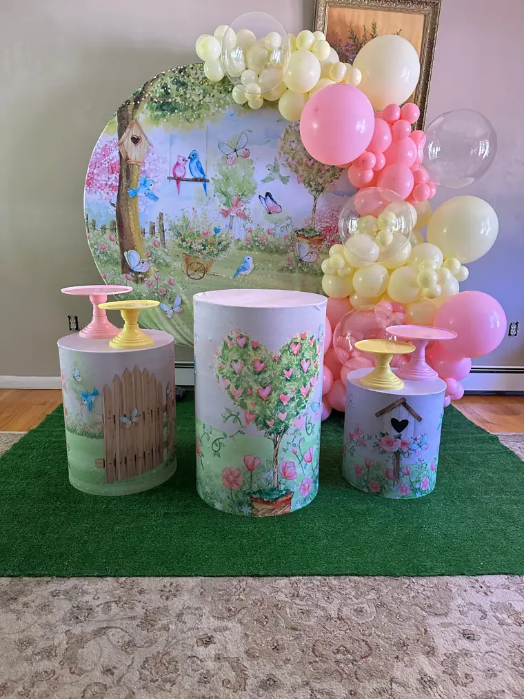 Decoração de jardim montada pela Party Decoration Fest: painel redondo pintado, balões amarelos e rosa e três cilindros com bandejas